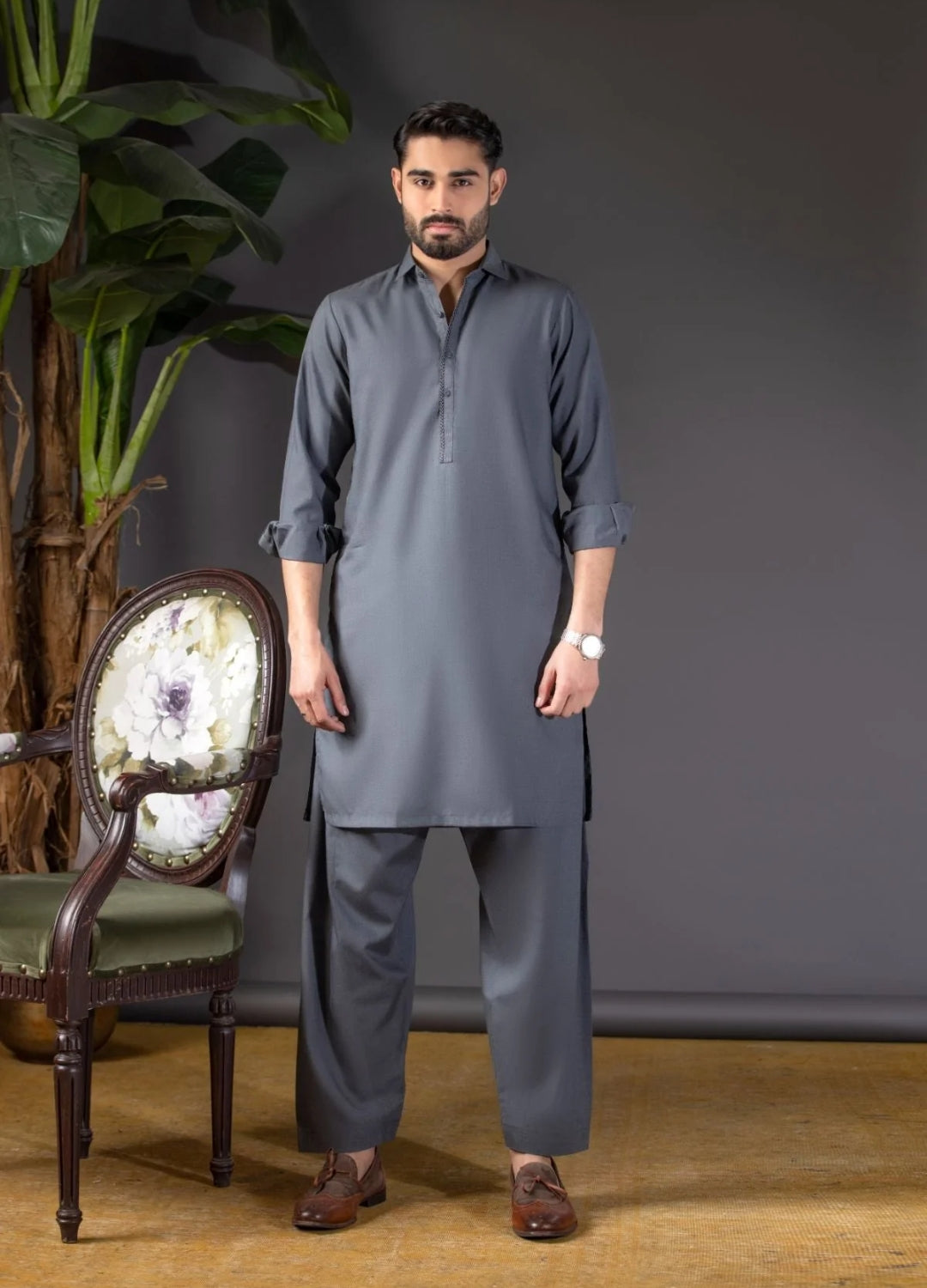 Man wearing a gray kurta with pants standing in a room with a chair and plant.