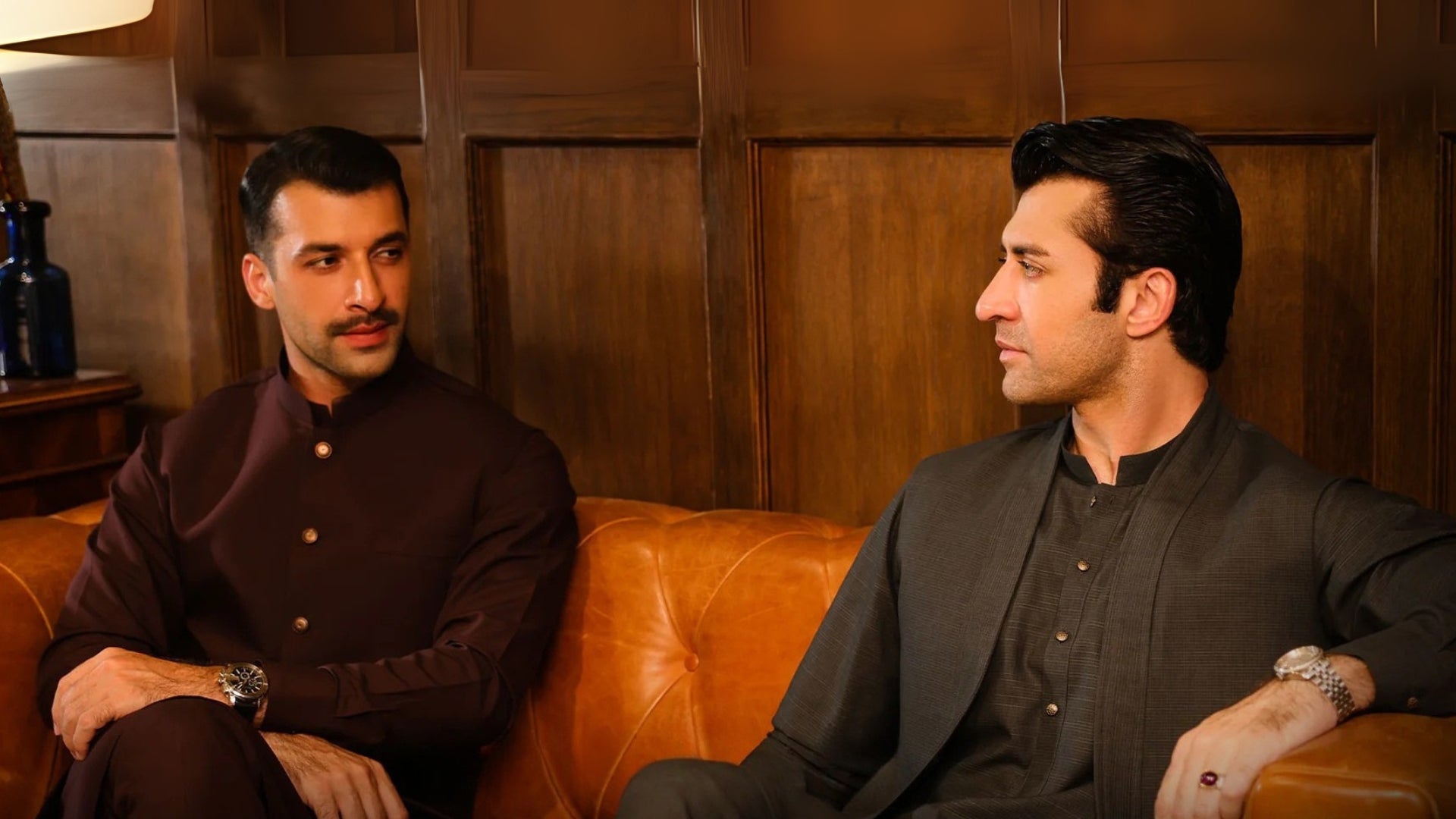 Two men sitting on a brown leather couch against a wooden paneled wall.